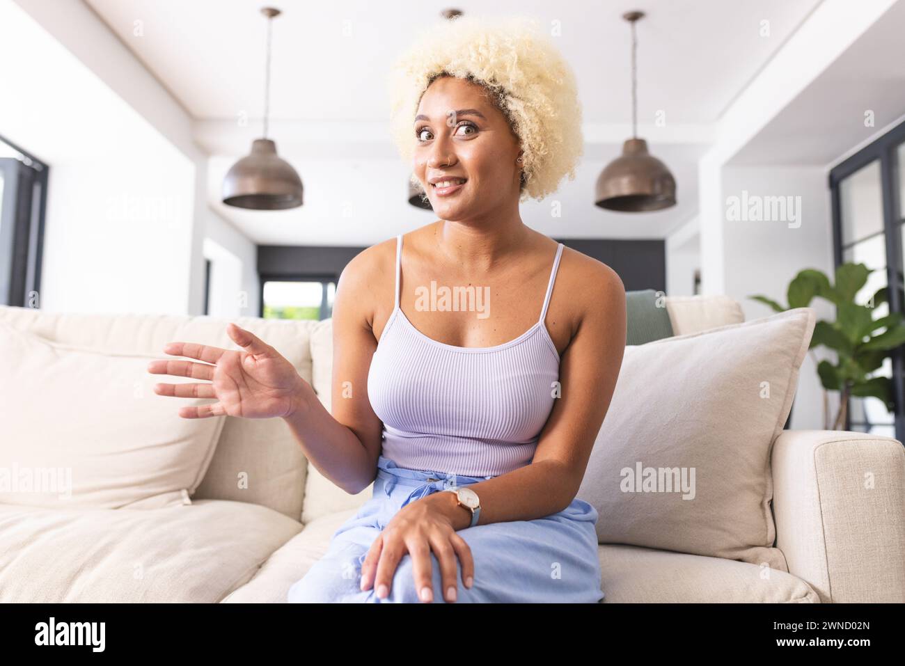 Giovane donna birazziale con capelli biondi ricci seduta sul divano, gestendo durante una videochiamata Foto Stock