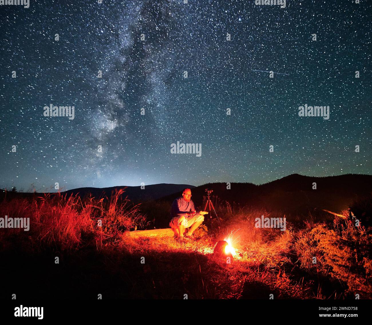 Uomo che scatta foto di cielo stellato con la via Lattea. Fotografo di sesso maschile seduto sul tronco vicino alla macchina fotografica. Uomo con macchina fotografica seduto accanto al fuoco del campo in montagna. Foto Stock