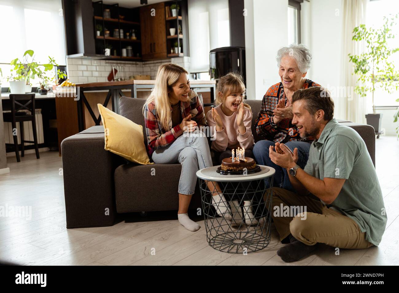 Una scena emozionante si svolge mentre una famiglia multigenerazionale si riunisce su un divano per presentare una torta di compleanno a una nonna adorata, creando ricordi indimenticabili Foto Stock