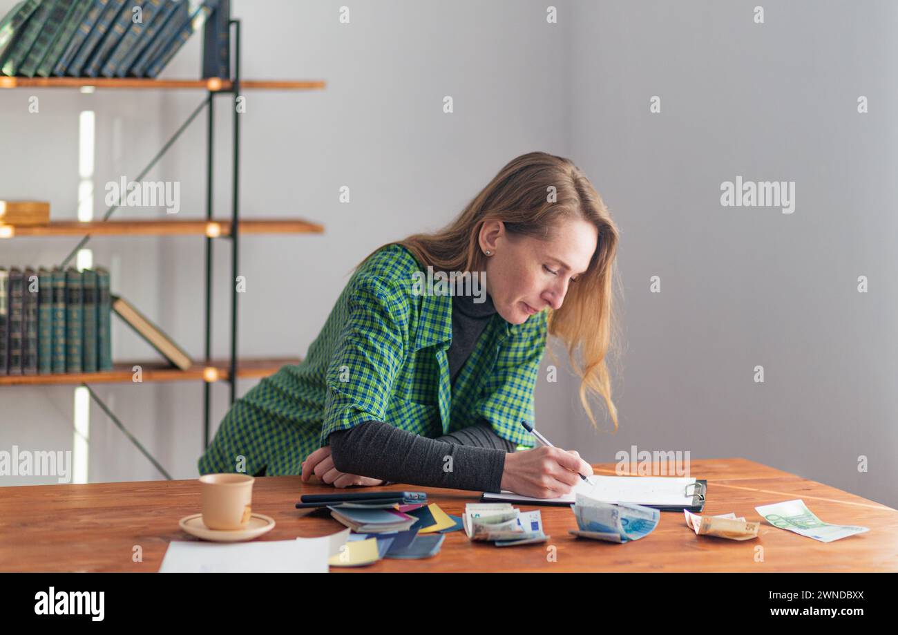 Una giovane bella signora prende appunti su un tablet sullo sfondo di un ambiente di lavoro e i soldi sono disposti sul tavolo. Foto Stock