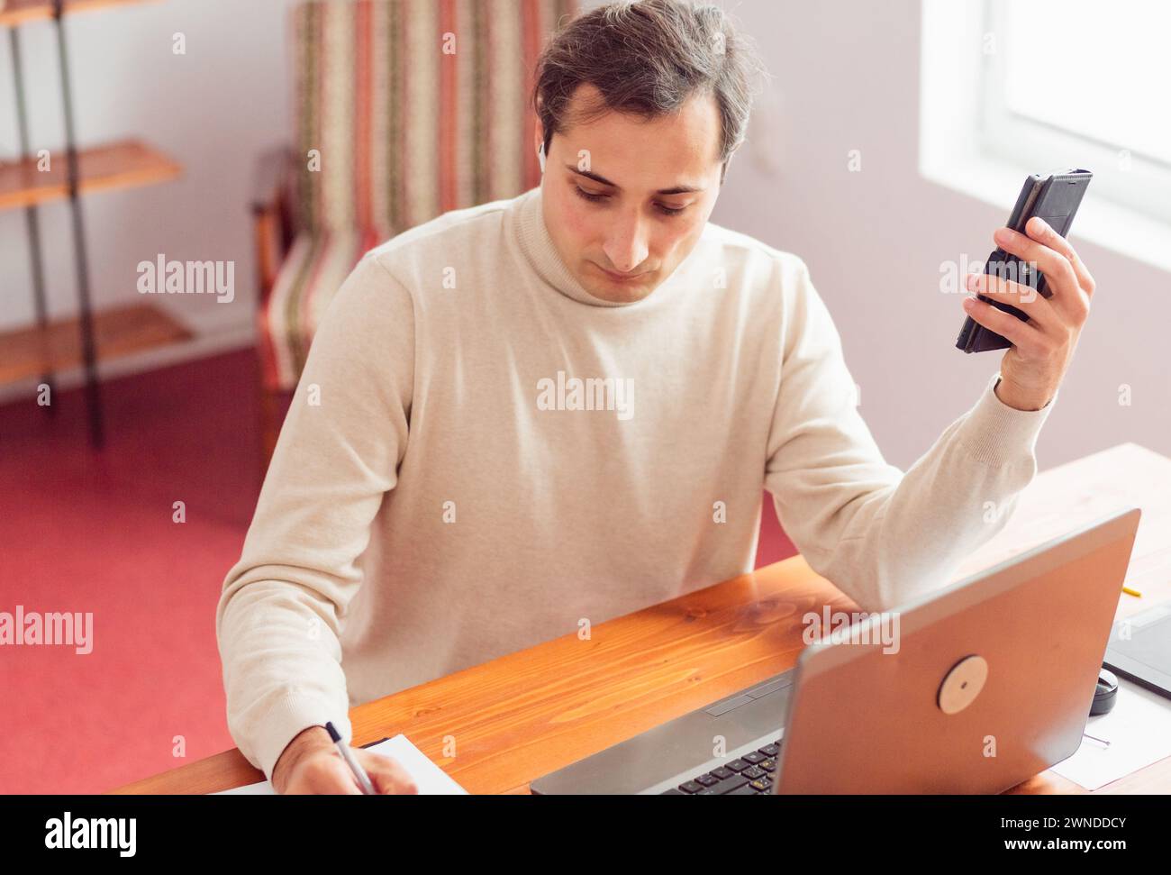 Giovane uomo adulto in ufficio a casa che lavora con un computer portatile e un telefono cellulare. Sul tavolo sono presenti tavolette grafiche elettroniche e cartacee. Foto Stock