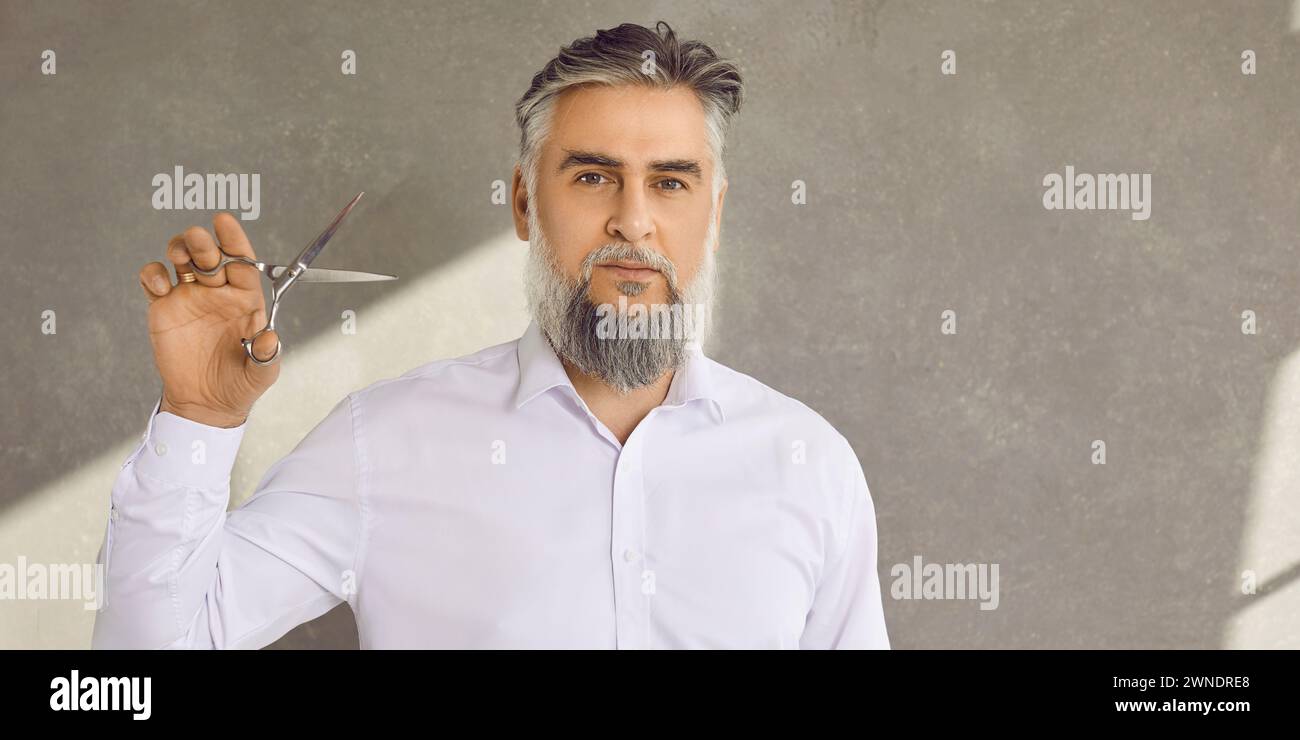 Ritratto di barbiere barbuto e baciato con forbici su sfondo grigio Foto Stock