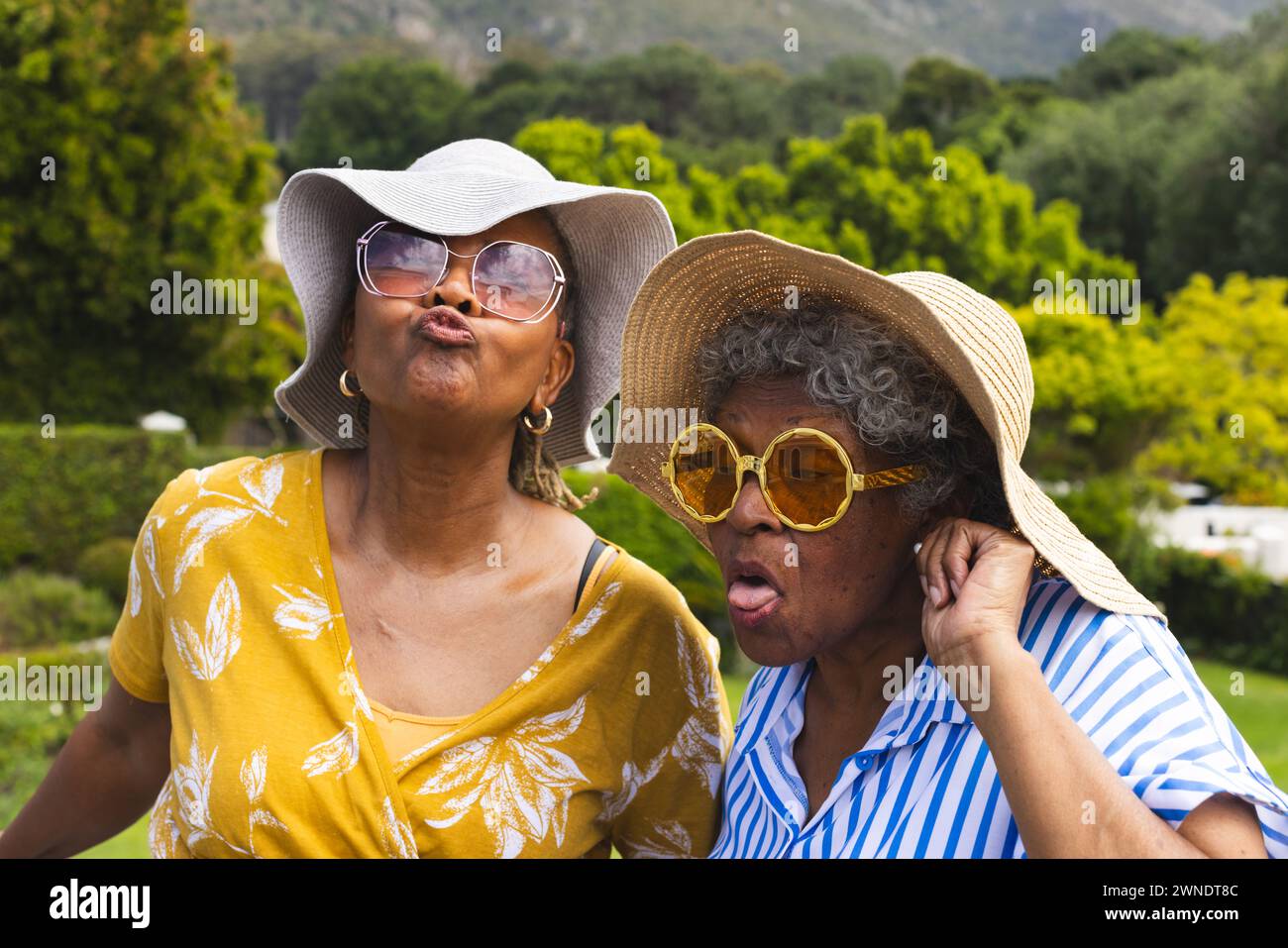 Donna afroamericana anziana e donna birazziale fanno facce giocose, indossando cappelli da sole, in vacanza Foto Stock