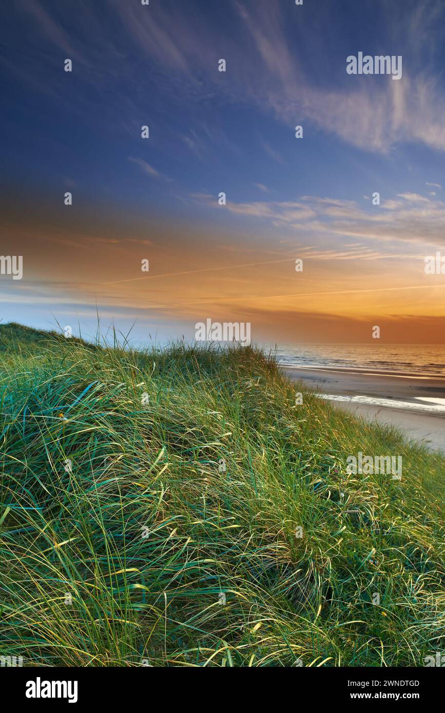 Dune erbose, tramonti e spiaggia nel paesaggio, nella natura e nell'ambiente all'aperto. Oceano, acqua e nuvole nel cielo blu con vista panoramica serale, sabbia e. Foto Stock