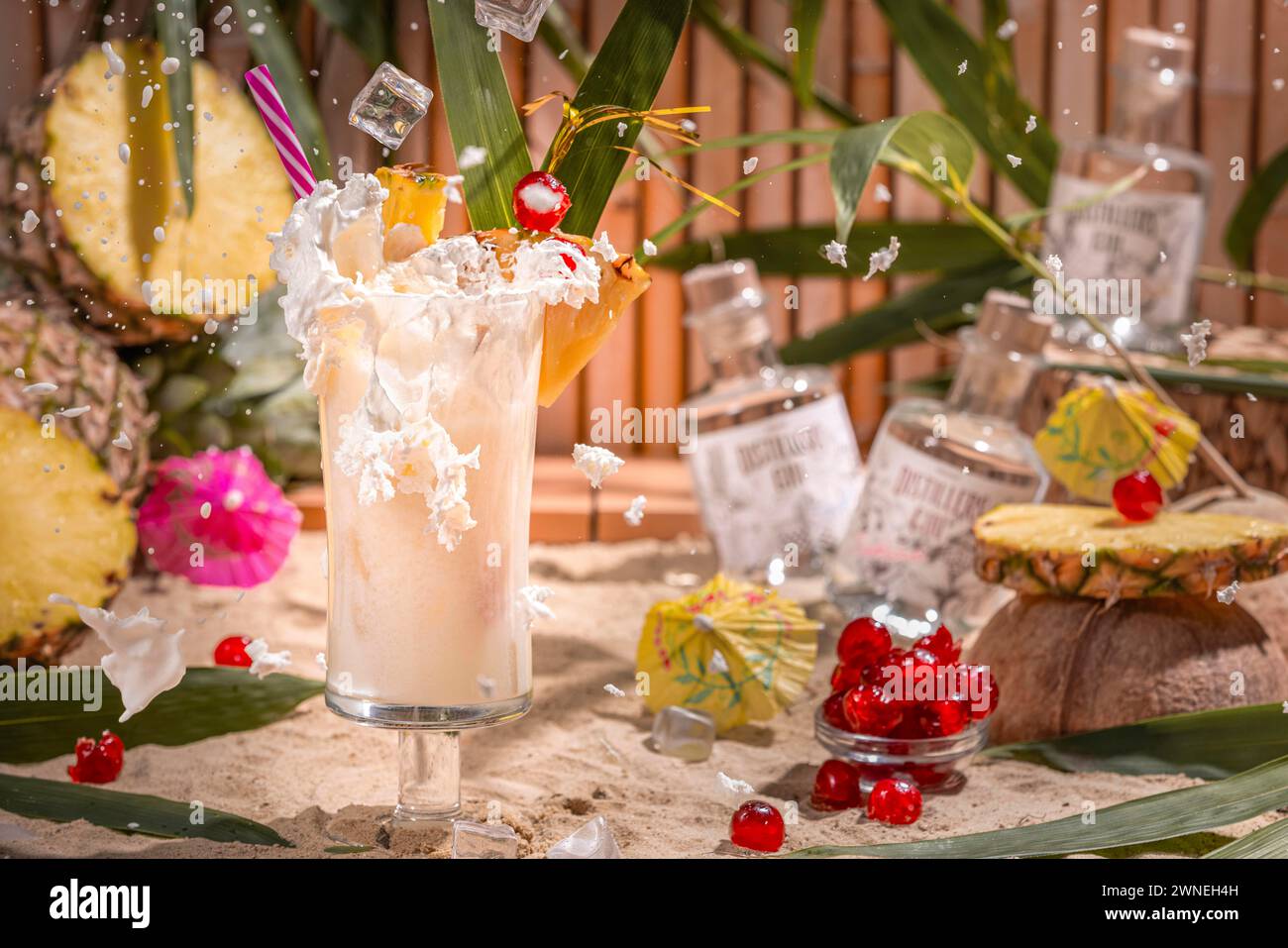 I cubetti di ghiaccio che cadono creano un panna spruzzante in un cocktail tropicale circondato da frutta, sabbia e bottiglie di gin Foto Stock