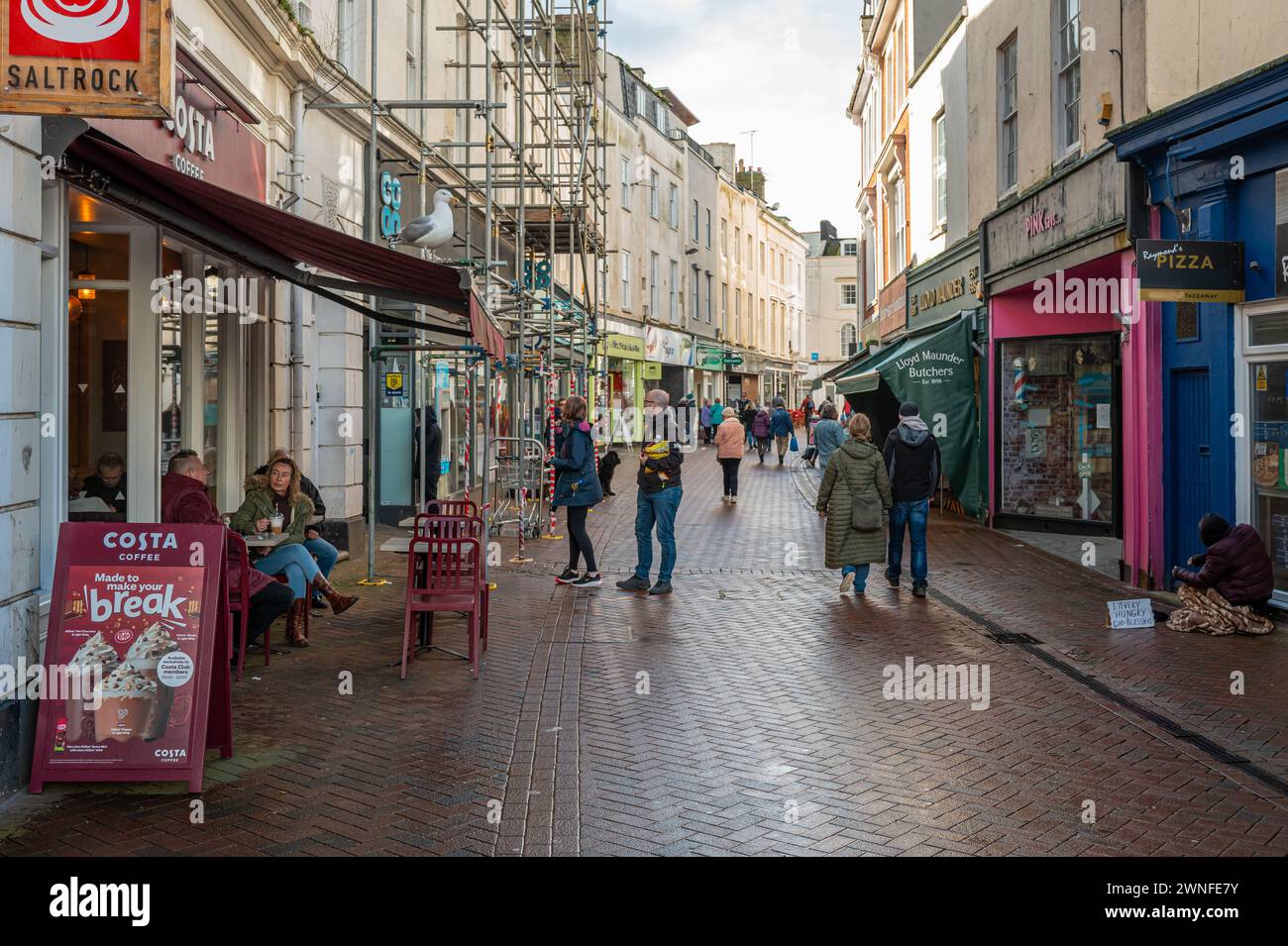 Via dello shopping nel centro di Teignmouth con un caffè Costa, pedoni e un senzatetto sul marciapiede. Foto Stock