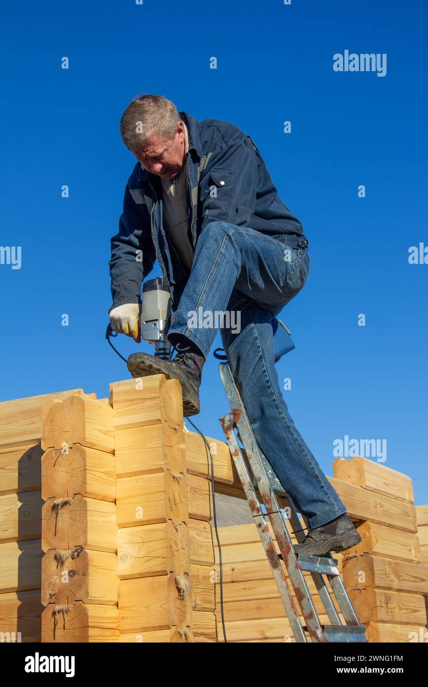 un uomo esegue un foro in una trave con un trapano, costruisce una casa in legno fatta di legno profilato su uno sfondo blu cielo Foto Stock
