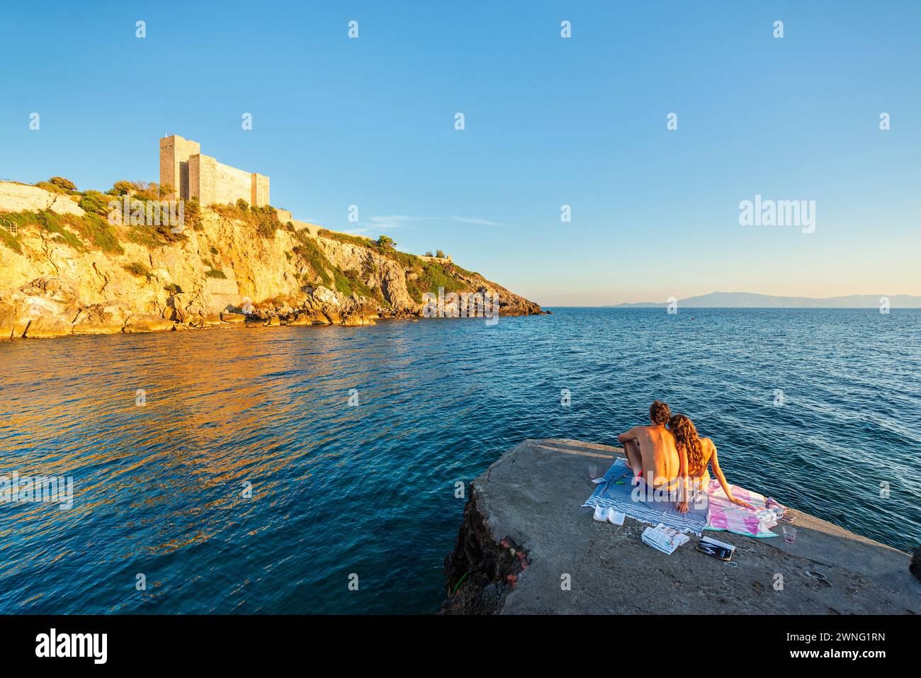 Una coppia amorevole che guarda il castello di Rocca Aldobrandesca sulla costa rocciosa della Maremma a Talamone nel tramonto dorato, Toscana, Italia Foto Stock