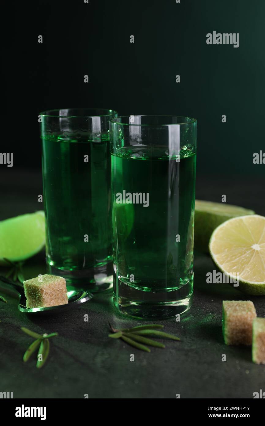 Absinthe in bicchieri da shot, cucchiaio, zucchero di canna, lime e rosmarino su un tavolo grigio. Bevanda alcolica Foto Stock