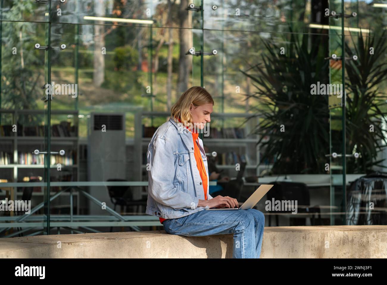 Un uomo concentrato con un laptop siede vicino a un edificio moderno e lavora come programmatore in un'azienda IT all'aperto. Foto Stock