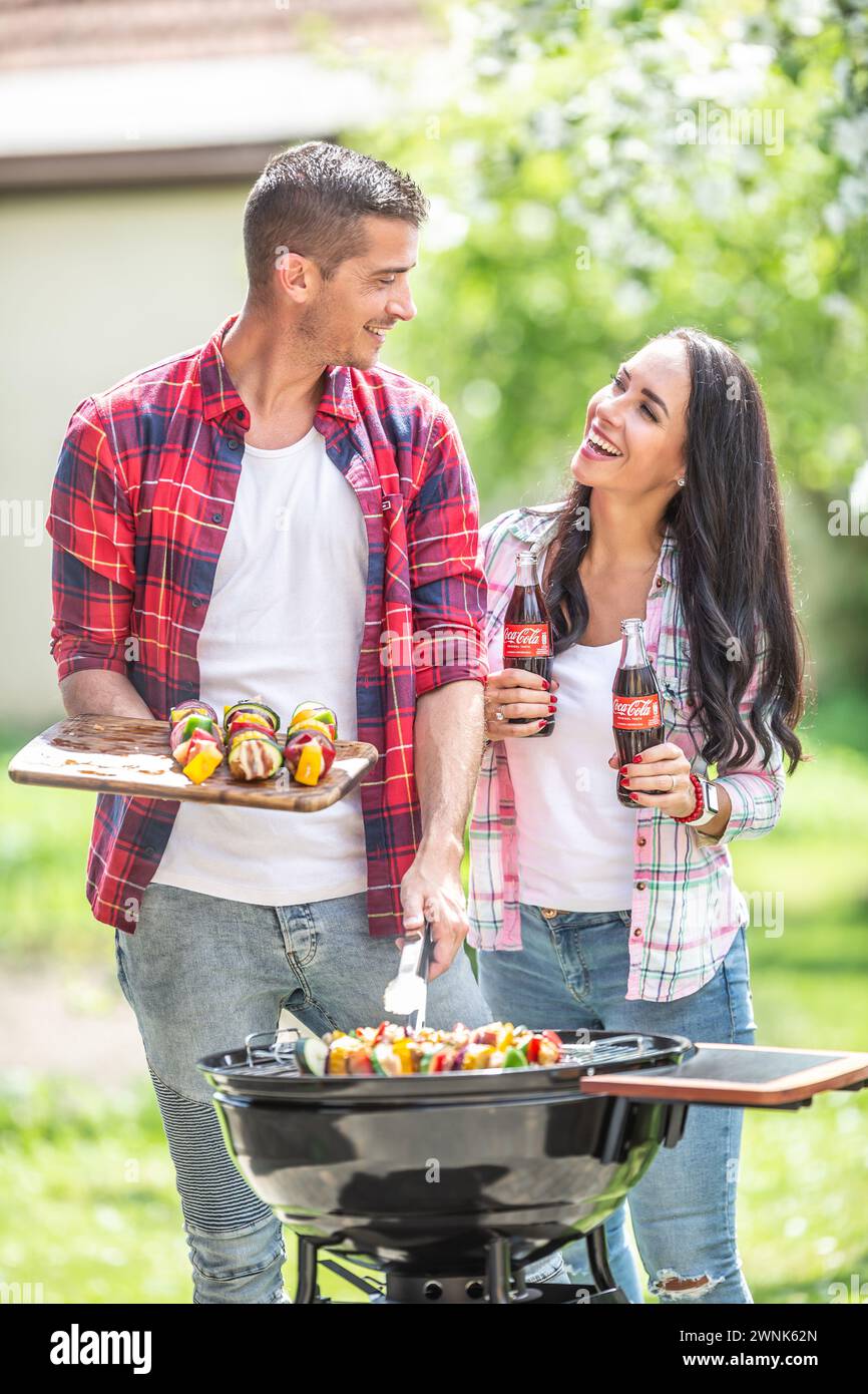 Marzo. 02. 2024 Liptovsky Mikulas. Slovacchia - la donna porta al marito una bottiglia di Coca Cola mentre sta barbecue sulla griglia in giardino. Foto Stock