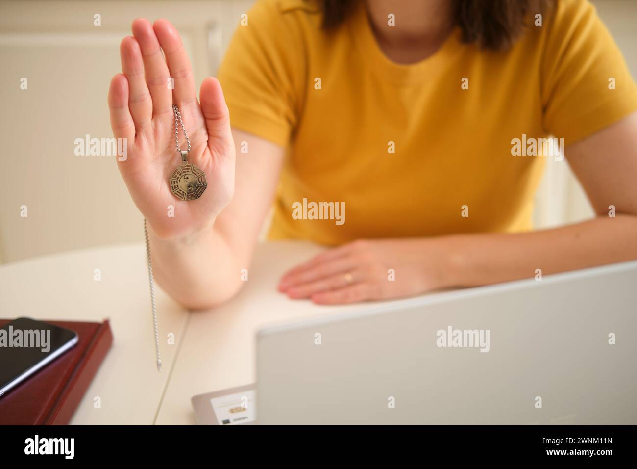 Una donna con un portatile tiene in mano un simbolo religioso buddista mentre è seduta a un tavolo in una cucina di casa. Una donna d'affari adulta lavora da casa, a r Foto Stock