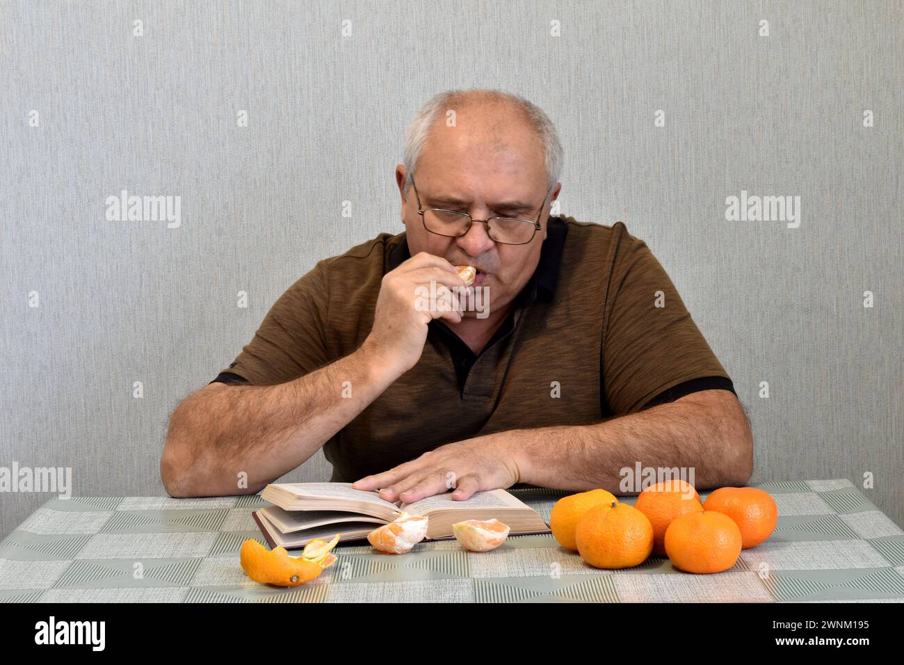 Un uomo si siede a un tavolo e legge un libro. Ci sono mandarini sul tavolo. Legge e mangia i mandarini. Foto Stock