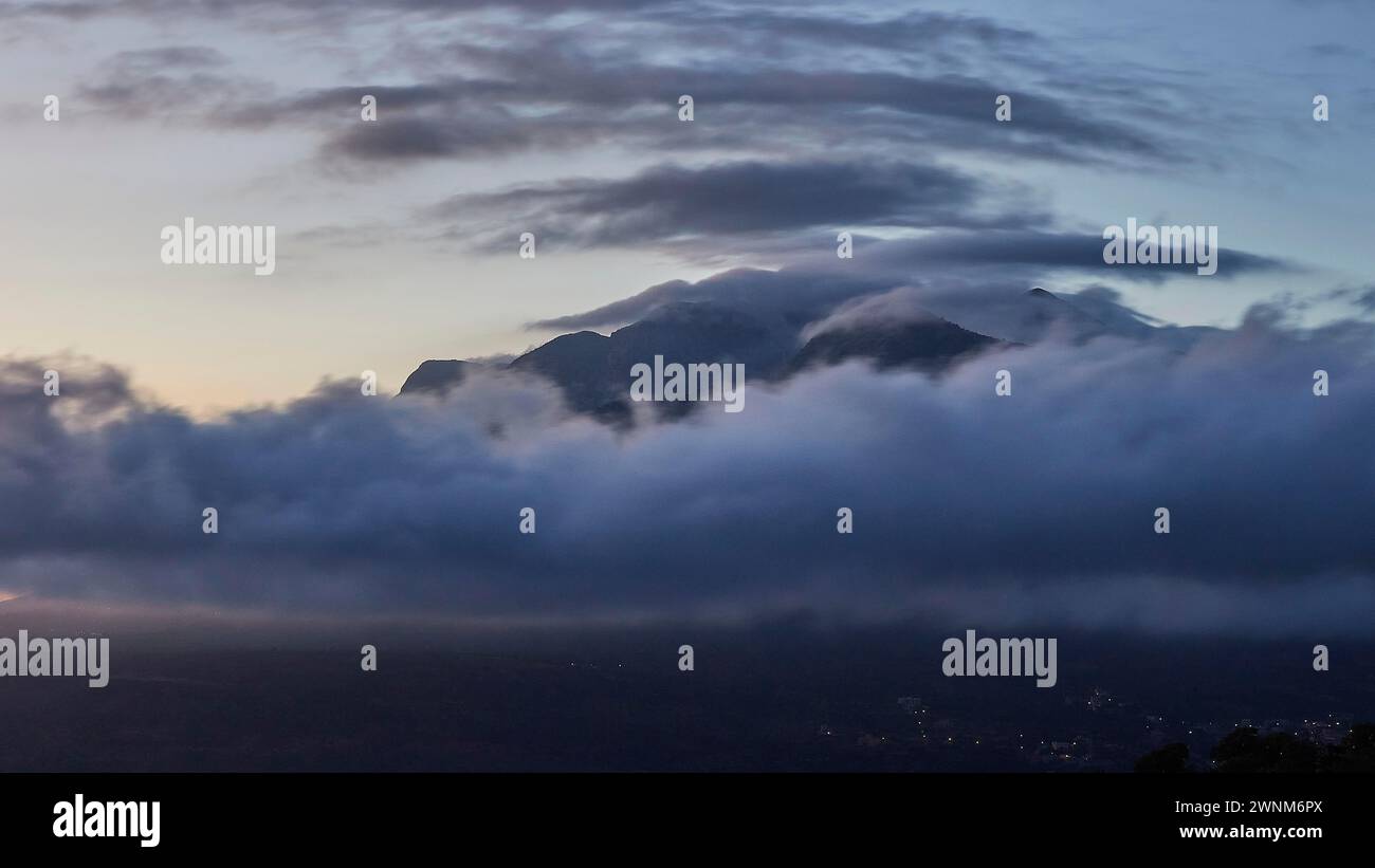 Scena mistica di un picco di montagna avvolto da nuvole al crepuscolo con le luci di una città nella valle, i monti Taygetos, la penisola di mani, il Peloponneso Foto Stock