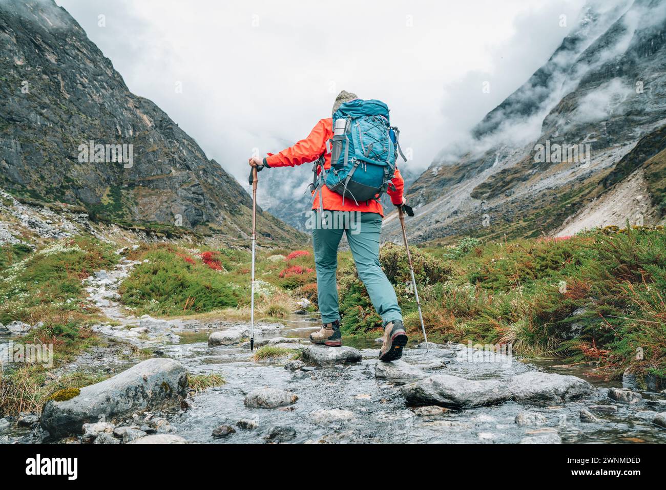 Moman con zaino e pali da trekking che attraversano il torrente di montagna durante il trekking nel Parco Nazionale di Makalu Barun in Nepal. Escursioni in montagna e persone attive conc Foto Stock