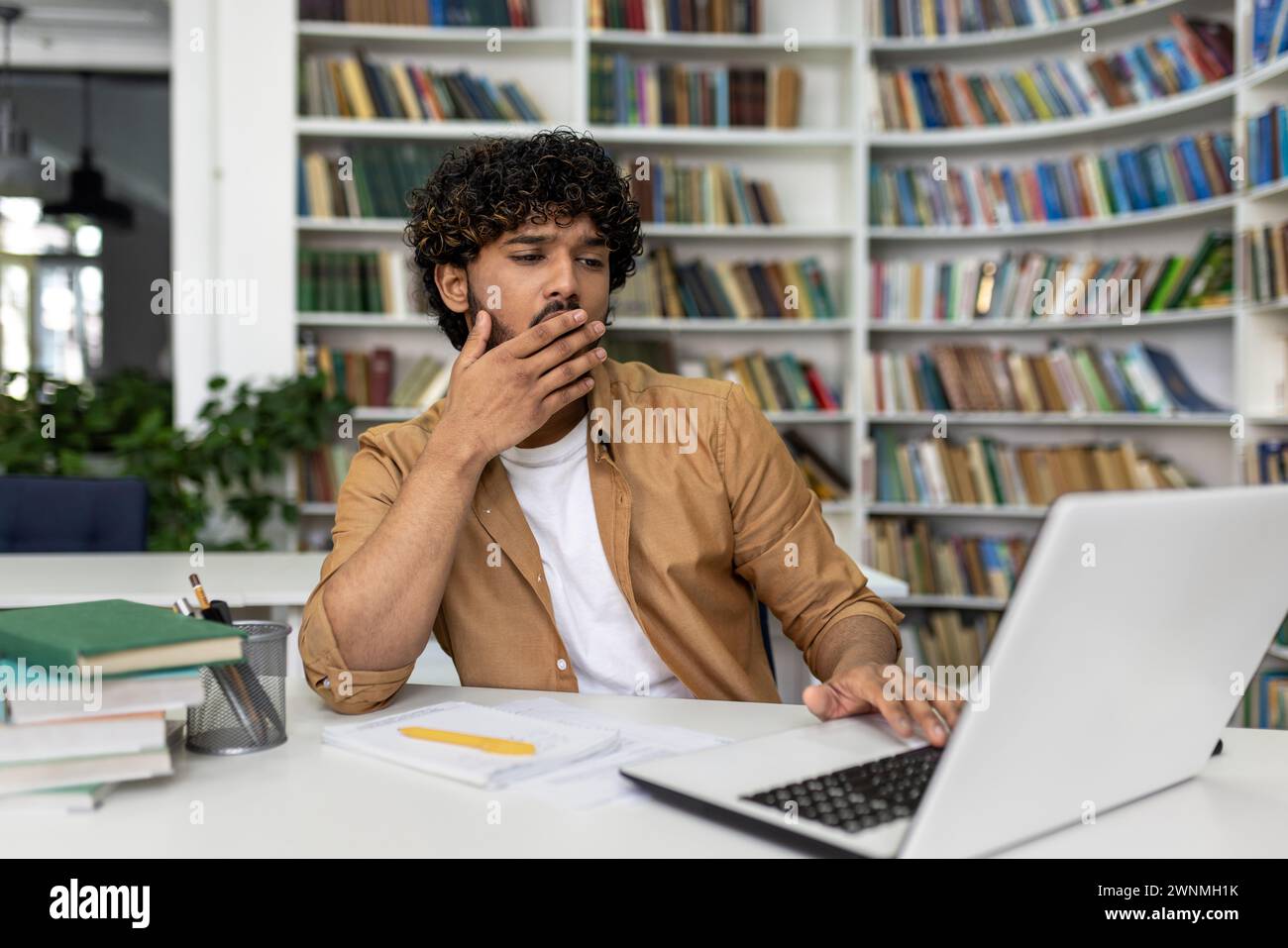 Uno studente maschile concentrato siede in una biblioteca, mano sul mento, profondamente pensato mentre studia sul suo portatile per gli esami futuri. I libri lo circondano, simboleggiando la ricerca accademica e l'apprendimento. Foto Stock