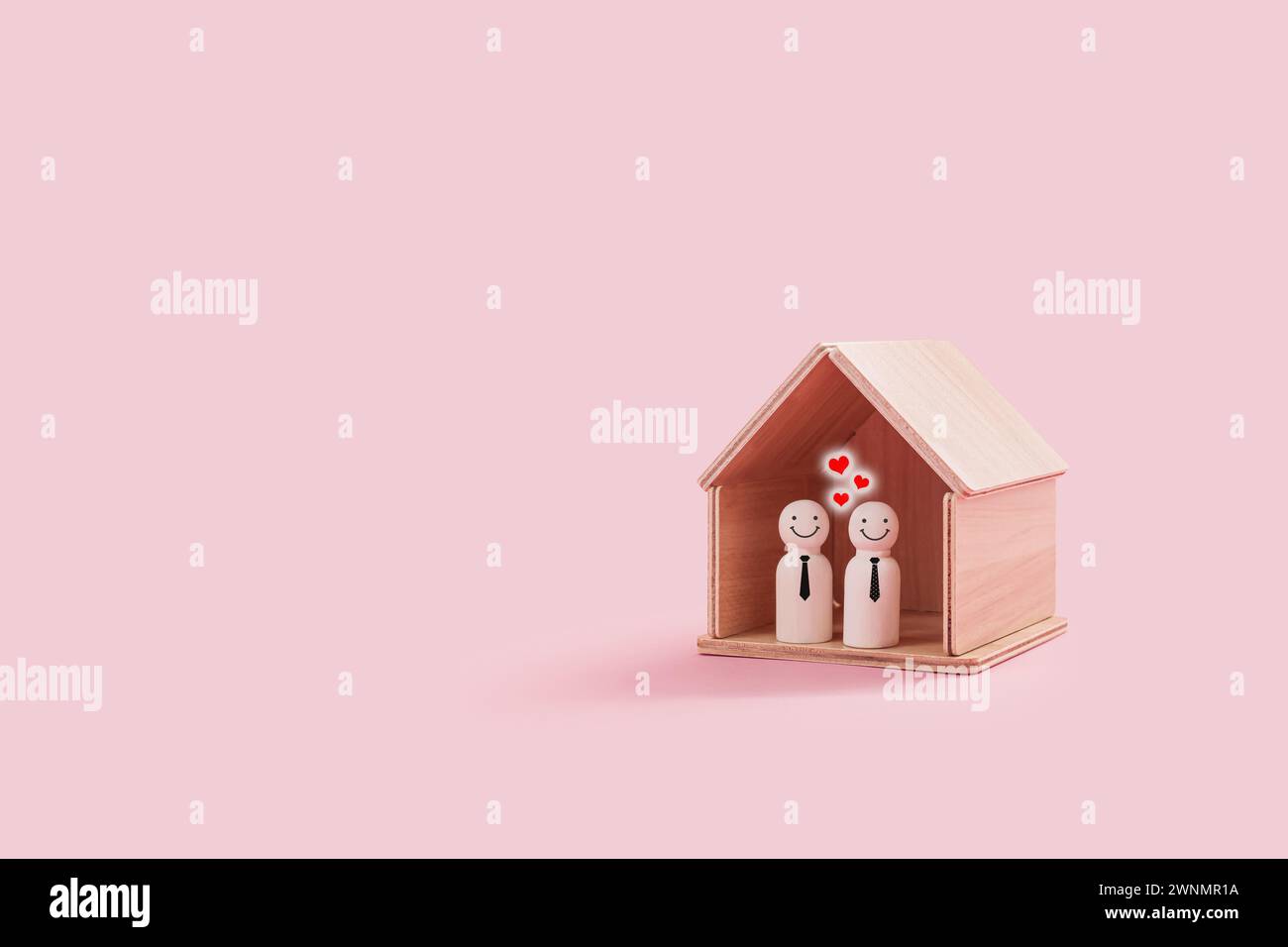 Figure di famiglia gay in legno felici all'interno di una piccola casa di legno isolata su uno sfondo rosa. Due maschi Pride sorridenti e vicini Foto Stock