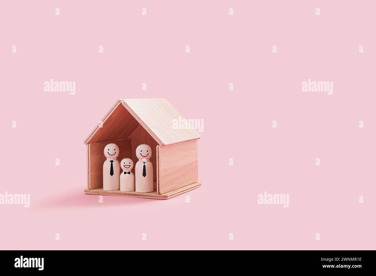 Figure di famiglia gay in legno felici all'interno di una piccola casa di legno isolata su uno sfondo rosa. Due padri dell'orgoglio e un ragazzo sorridente stanno vicini Foto Stock