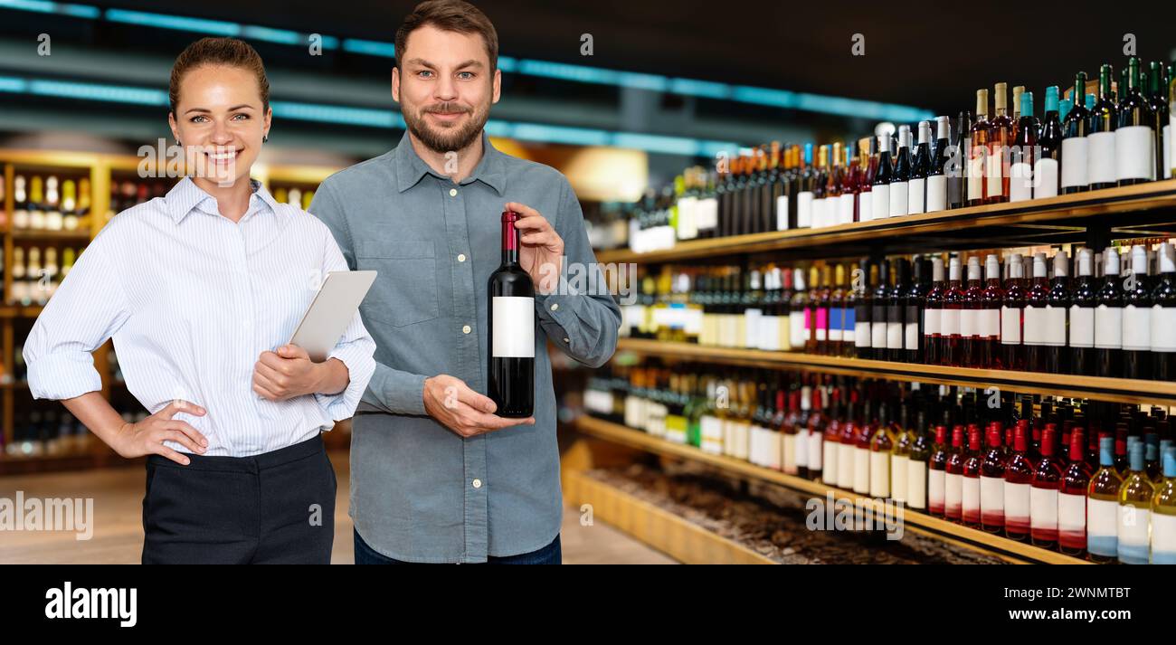 Concetto di rivenditore di alcolici. I proprietari d'affari, donna e uomo, sorridono davanti alle bottiglie di vino sugli scaffali del loro negozio. Foto Stock