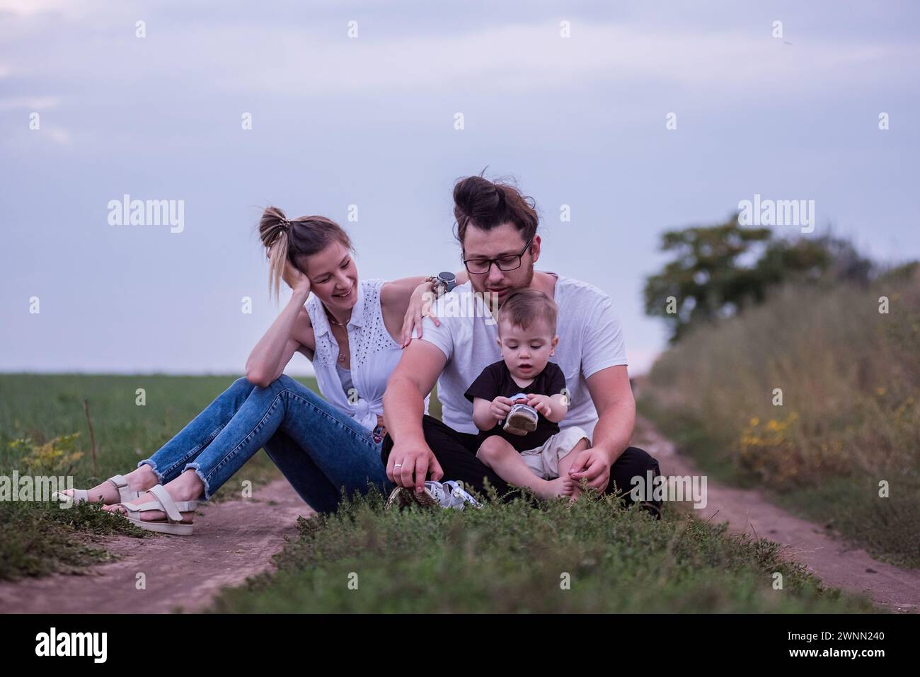La famiglia Diversity di tre persone gode di momenti tranquilli insieme, seduti lungo i sentieri di campagna in piena luce del giorno, immersi nella natura. Tempo in famiglia a piedi Foto Stock