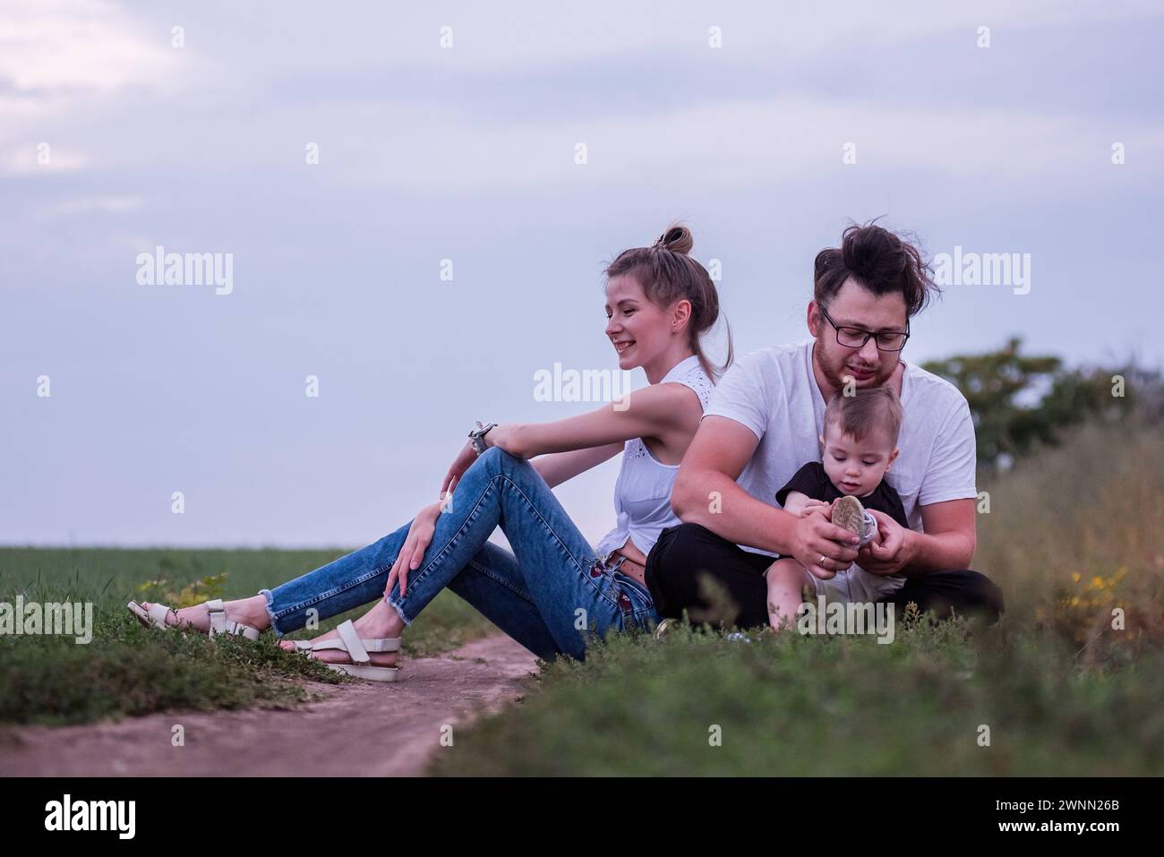 La famiglia Diversity di tre persone gode di momenti tranquilli insieme, seduti lungo i sentieri di campagna in piena luce del giorno, immersi nella natura. Tempo in famiglia a piedi Foto Stock