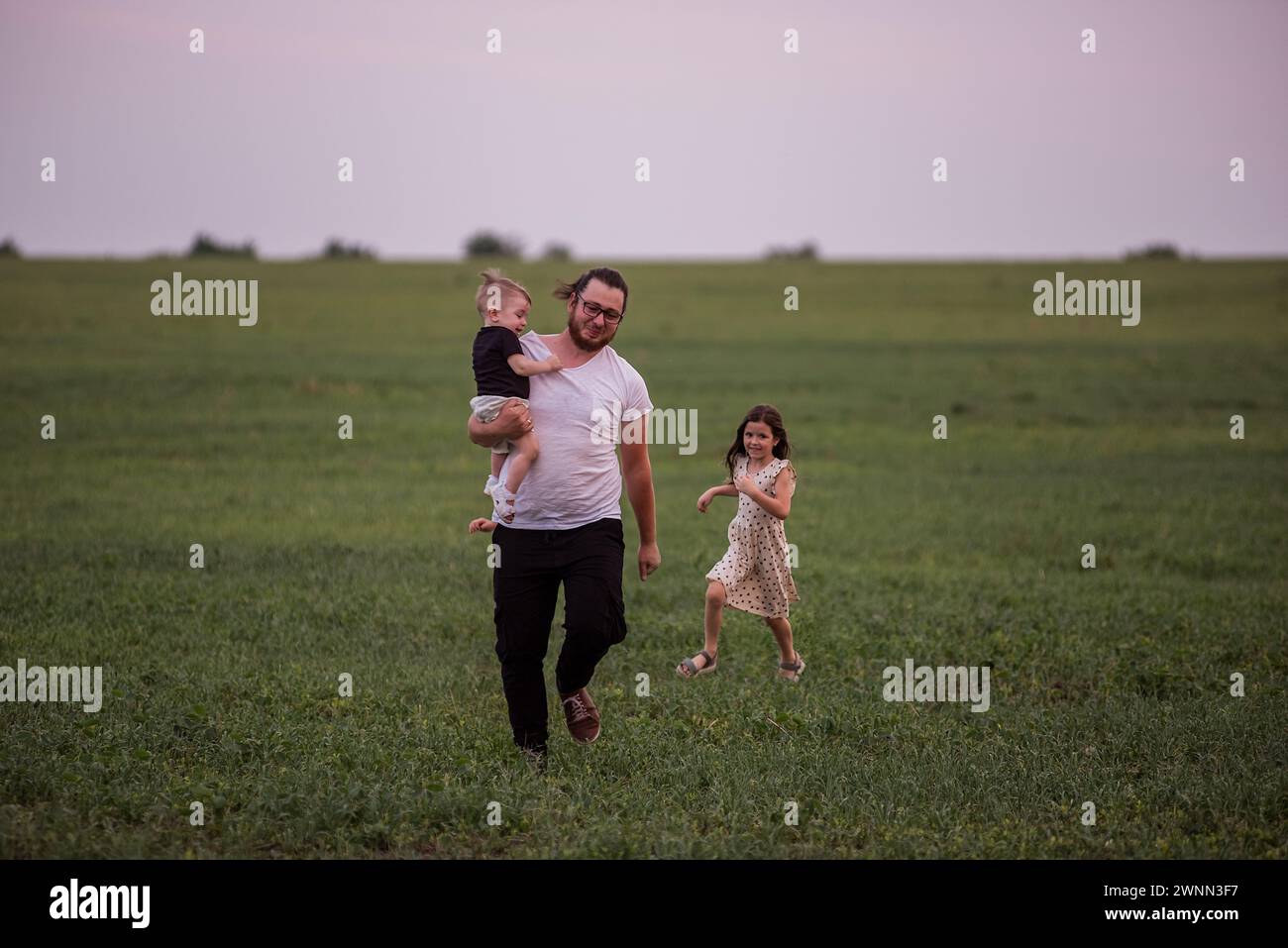 Il giovane padre corre con il figlio piccolo in braccio e la figlia maggiore attraverso il campo verde. Diversita' tempo giocoso in famiglia a Meadow. Festa dei padri. Foto Stock