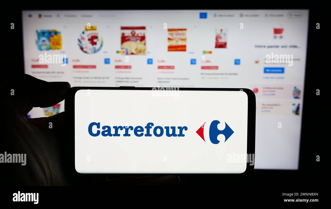 Persona che detiene un cellulare con il logo della società di vendita al dettaglio francese Carrefour SA di fronte alla pagina web aziendale. Mettere a fuoco il display del telefono. Foto Stock