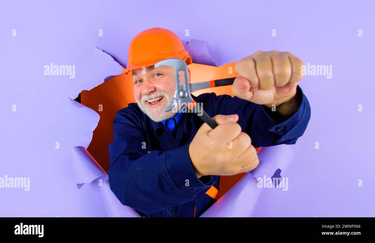 Operaio edile o costruttore in elmetto con chiave regolabile che guarda attraverso il foro della carta. Uomo con barba in uniforme con chiave regolabile Foto Stock