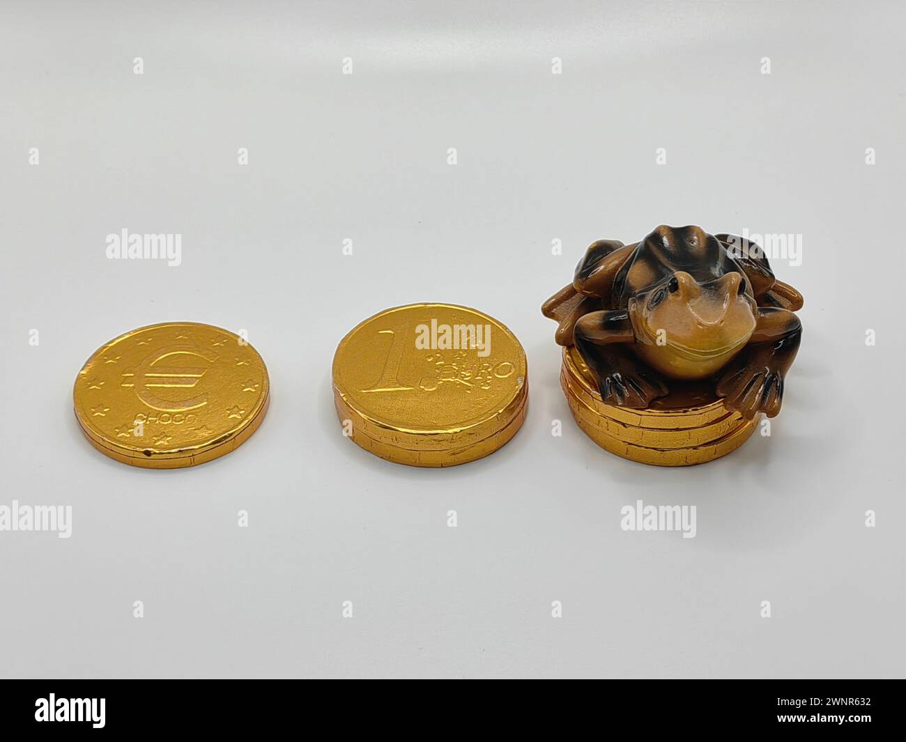 Rana nera e marrone giocattolo in ceramica, seduta sulle monete, cocco avvolge dolci. Testa o croce. Concetto di economia. Sfondo white paper. Foto Stock