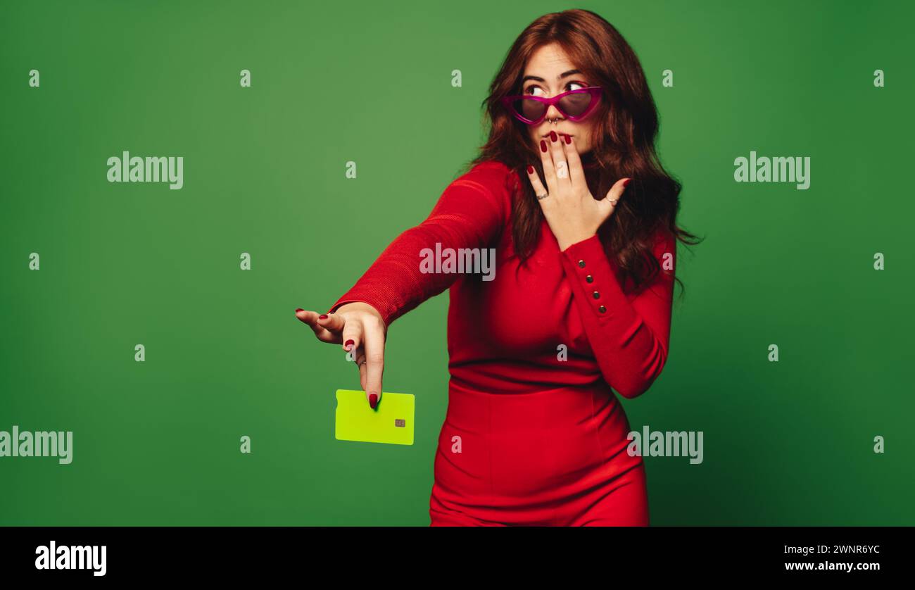 Una giovane donna con eleganti occhiali da sole regge una carta di credito verde su uno sfondo verde. E' pronta a pagare senza contatto per il suo trendy f Foto Stock