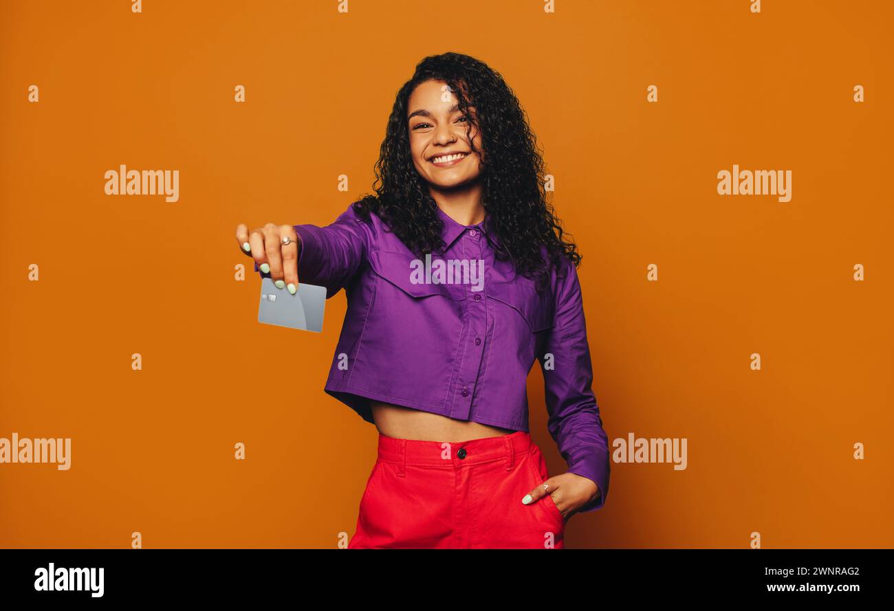 Una donna ha in mano una carta di credito, sorridendo felicemente su sfondo arancione. Dimostra il pagamento elettronico contactless toccando la carta. Alla moda Foto Stock