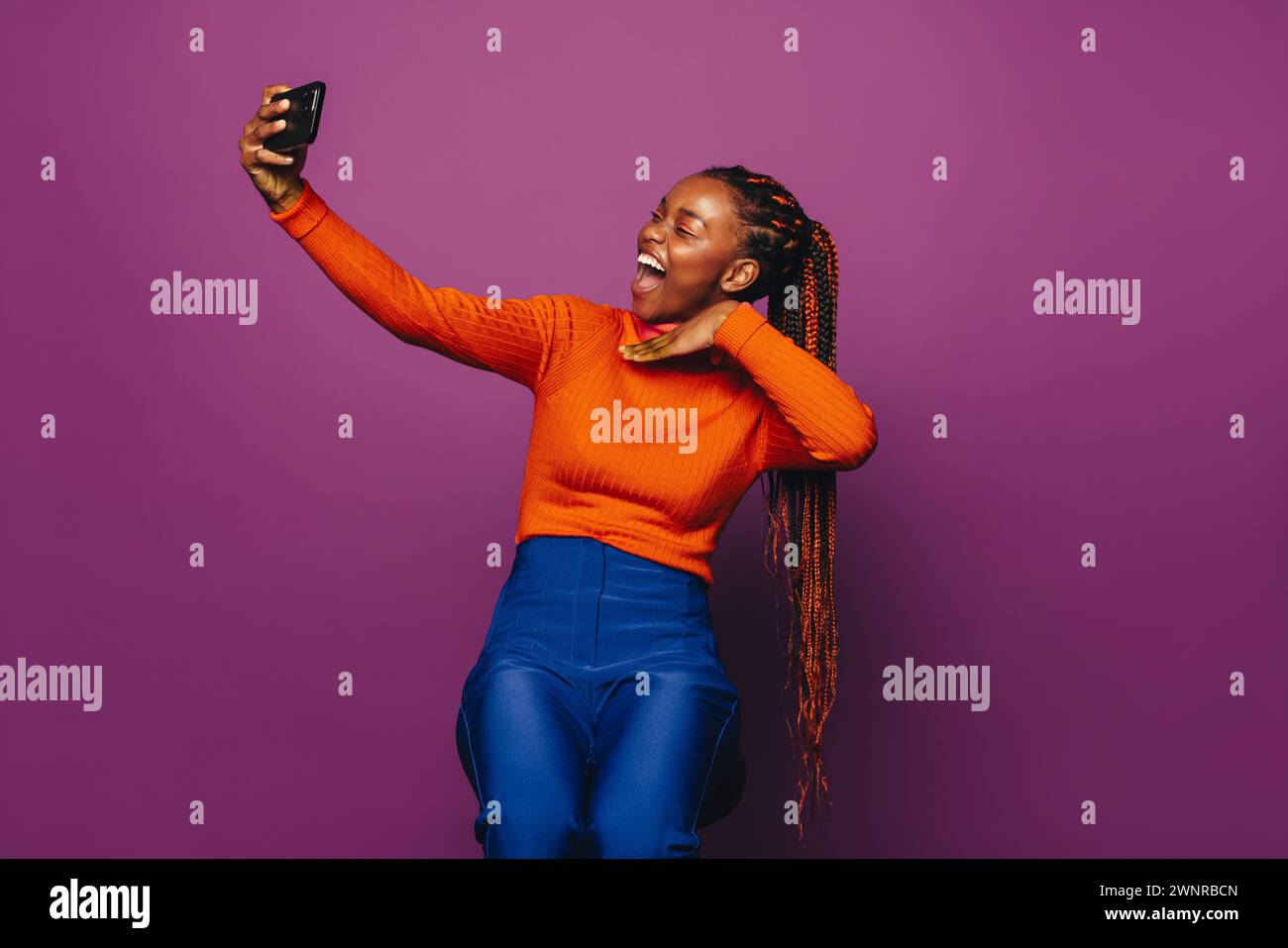 Gen Z Girl scatta un selfie vibrante con uno smartphone su uno sfondo viola colorato. Il suo abbigliamento casual e le trecce bicolore riflettono la sua vita moderna Foto Stock