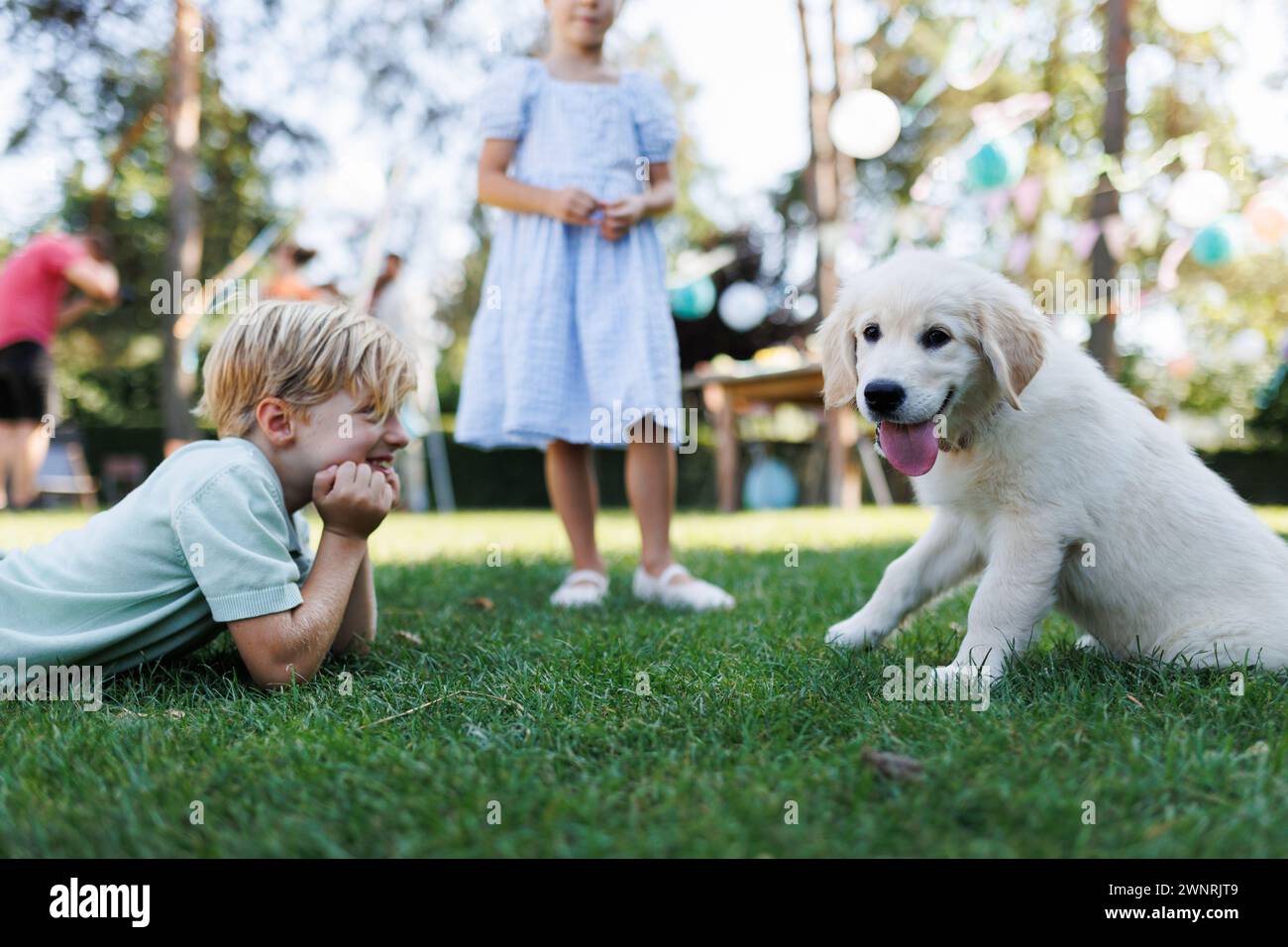 Bambini che giocano con un piccolo cucciolo alla festa in giardino di famiglia. Ritratto di un ragazzino steso sull'erba che guarda il cucciolo Golden retriever. Foto Stock