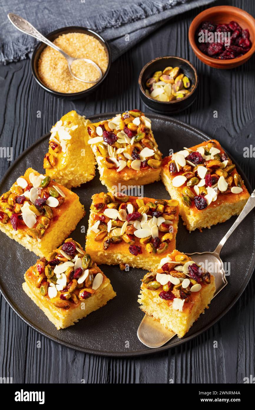 fette di torta, torta allo yogurt ricoperta di pistacchio, fette di mandorle e mirtilli rossi secchi su piatto nero con pala per torta su un tavolo di legno nero, Foto Stock
