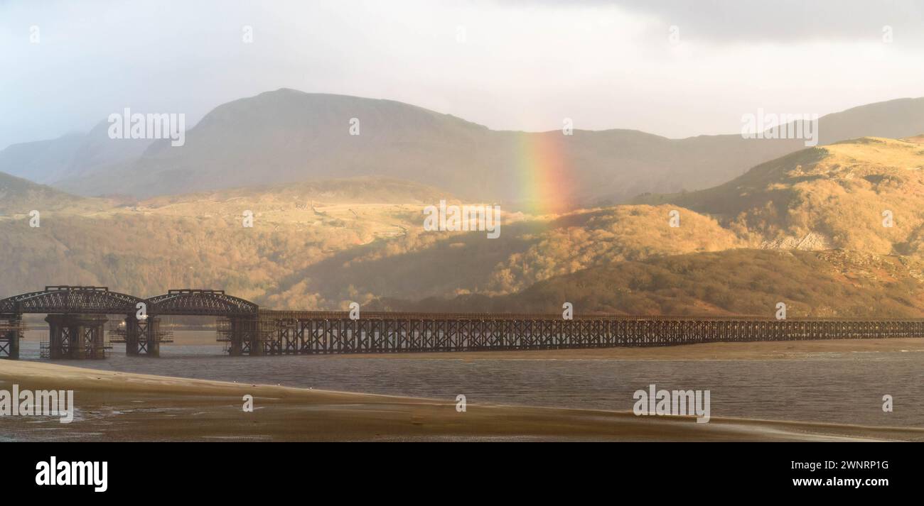 Un raggio di sole attraversa le nuvole di tempesta creando un arcobaleno sopra Barmouth Bridge (Pont Abermaw) Gwynedd Wales UK. Marzo 2024 Foto Stock