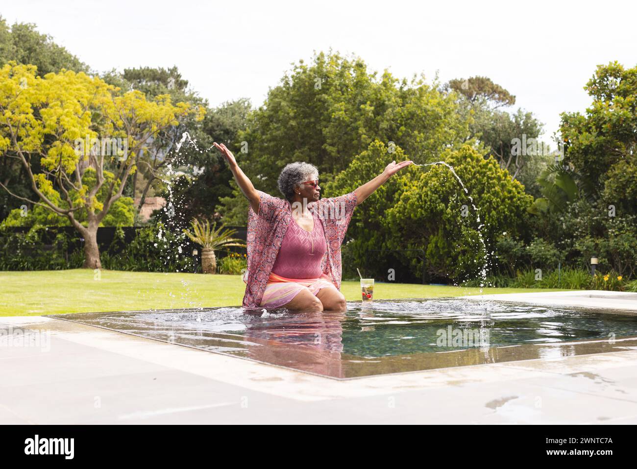 Una donna birazziale senior si tuffa gioiosamente in piscina, beve in mano, in mezzo al verde. Foto Stock