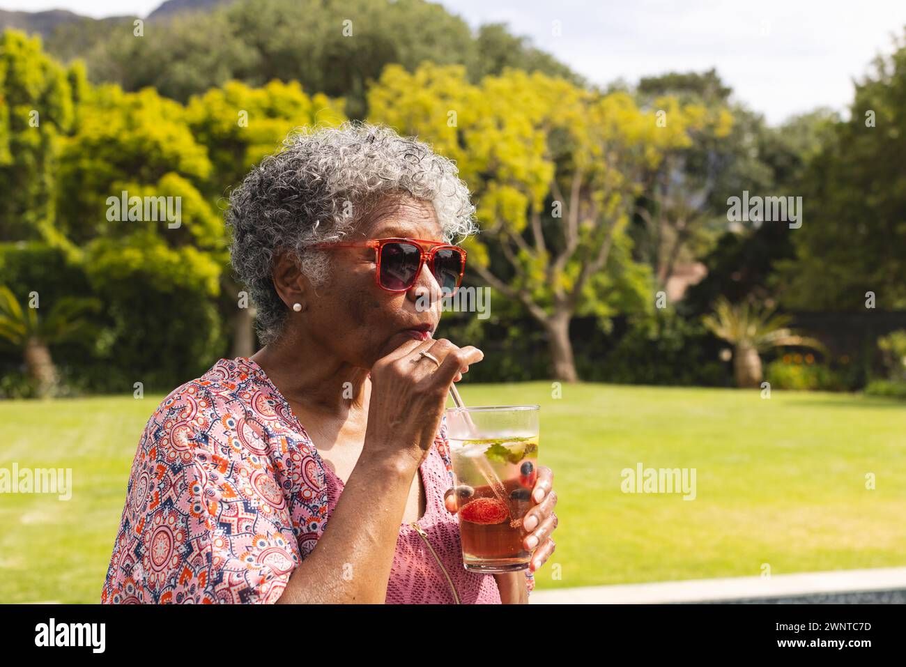 Una donna birazziale senior si gode un drink all'aperto, indossando occhiali da sole rossi e una camicetta a motivi geometrici Foto Stock