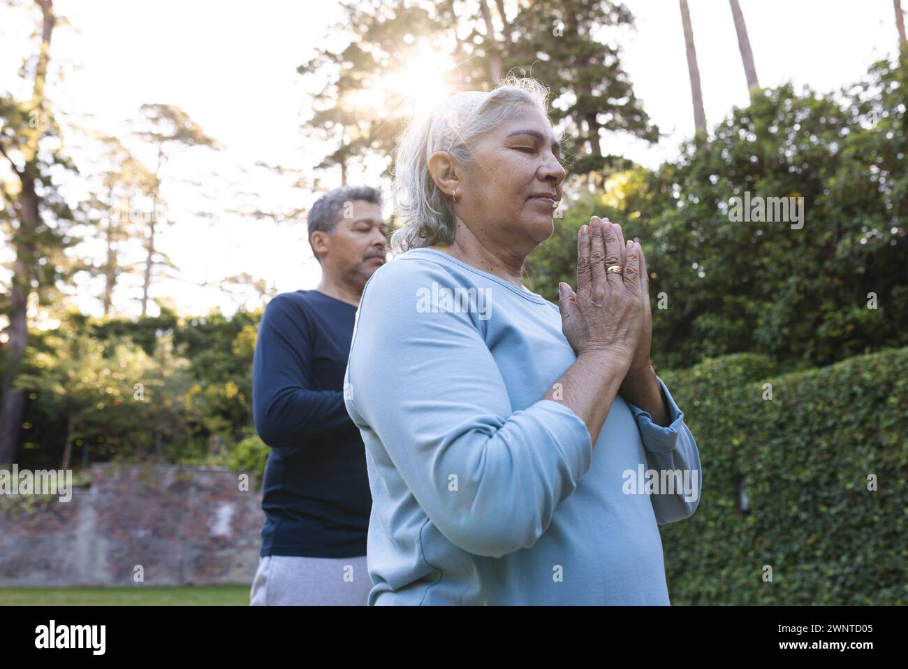 Donna e uomo birazziali senior meditano all'aperto a casa, filtrando la luce del sole attraverso gli alberi Foto Stock