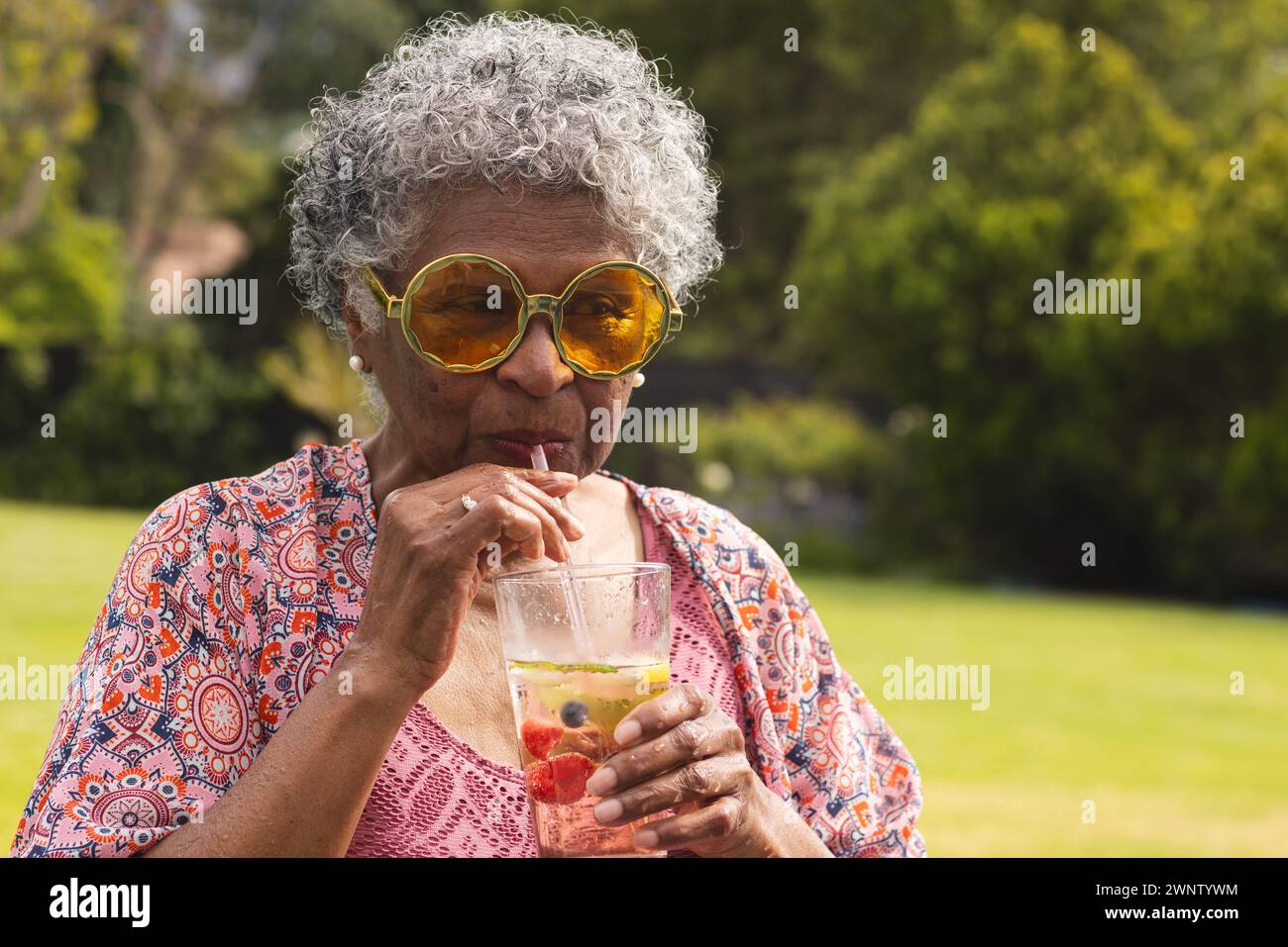 Una donna birazziale anziana con capelli ricci grigi sorseggia un drink attraverso una cannuccia Foto Stock