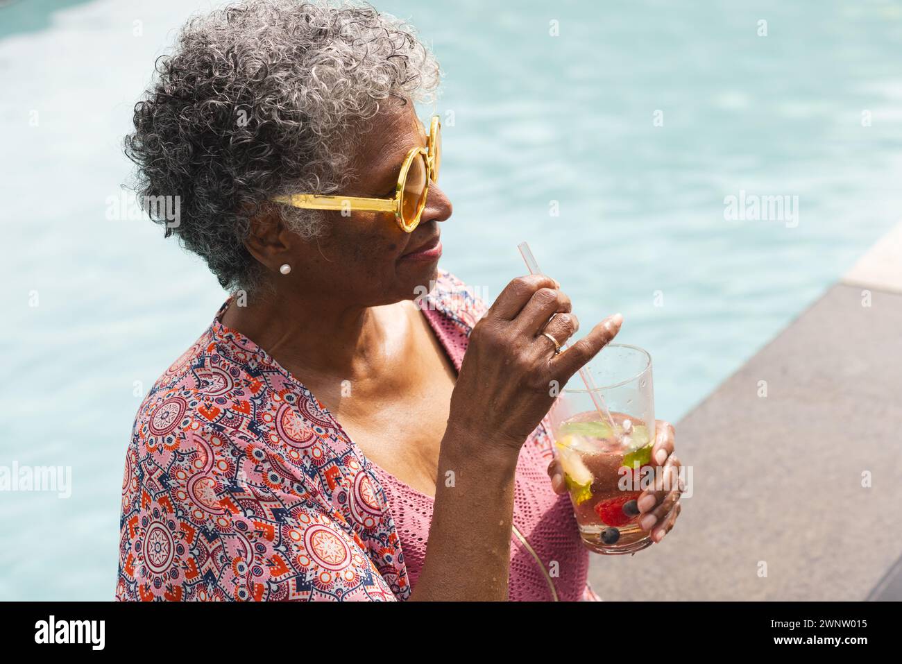 Una donna birazziale senior si gode un drink in piscina, indossando occhiali da sole gialli Foto Stock