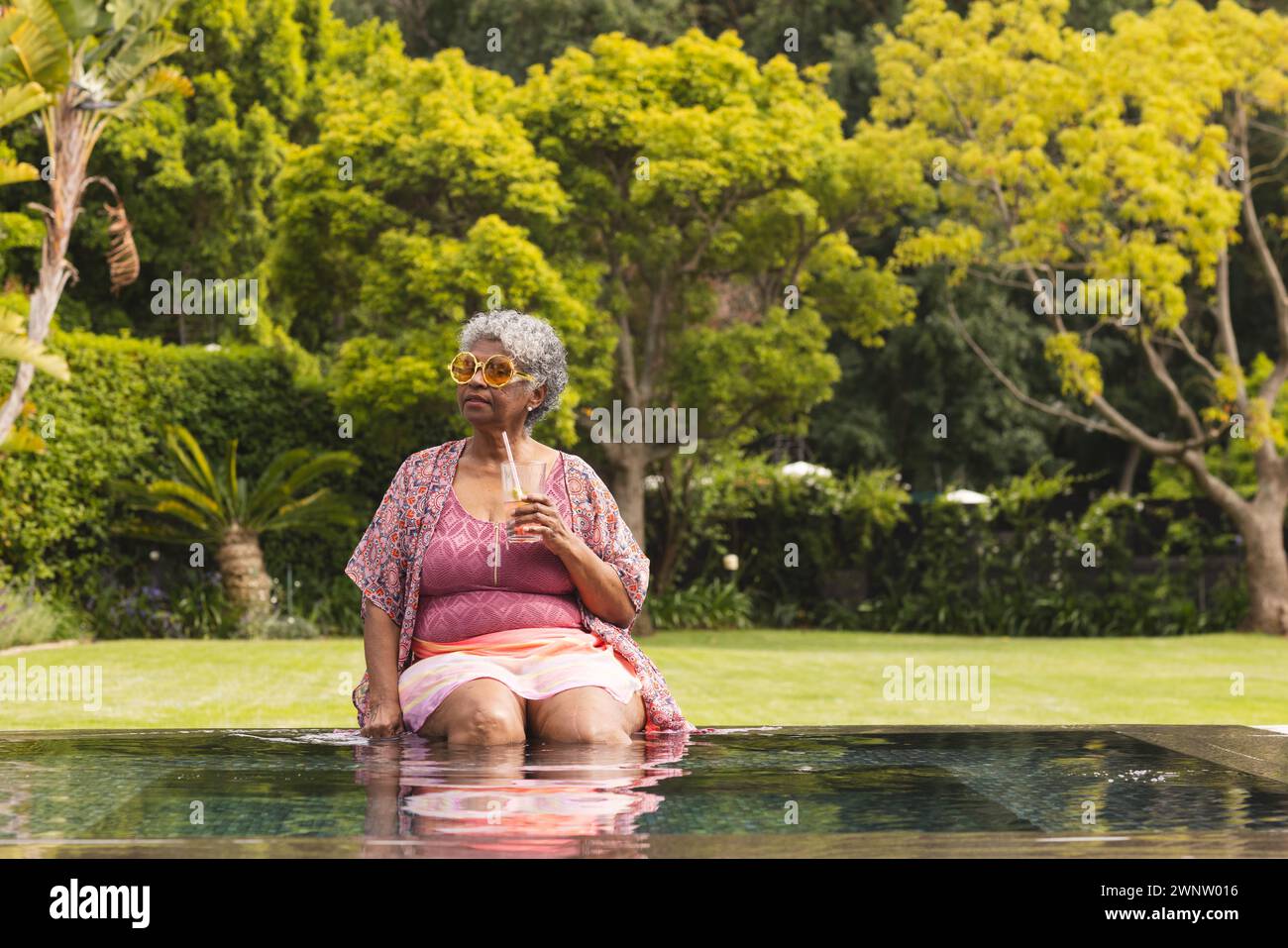 Donna birazziale senior con capelli grigi si rilassa in piscina, tenendo in mano un bicchiere, con spazio per le copie Foto Stock