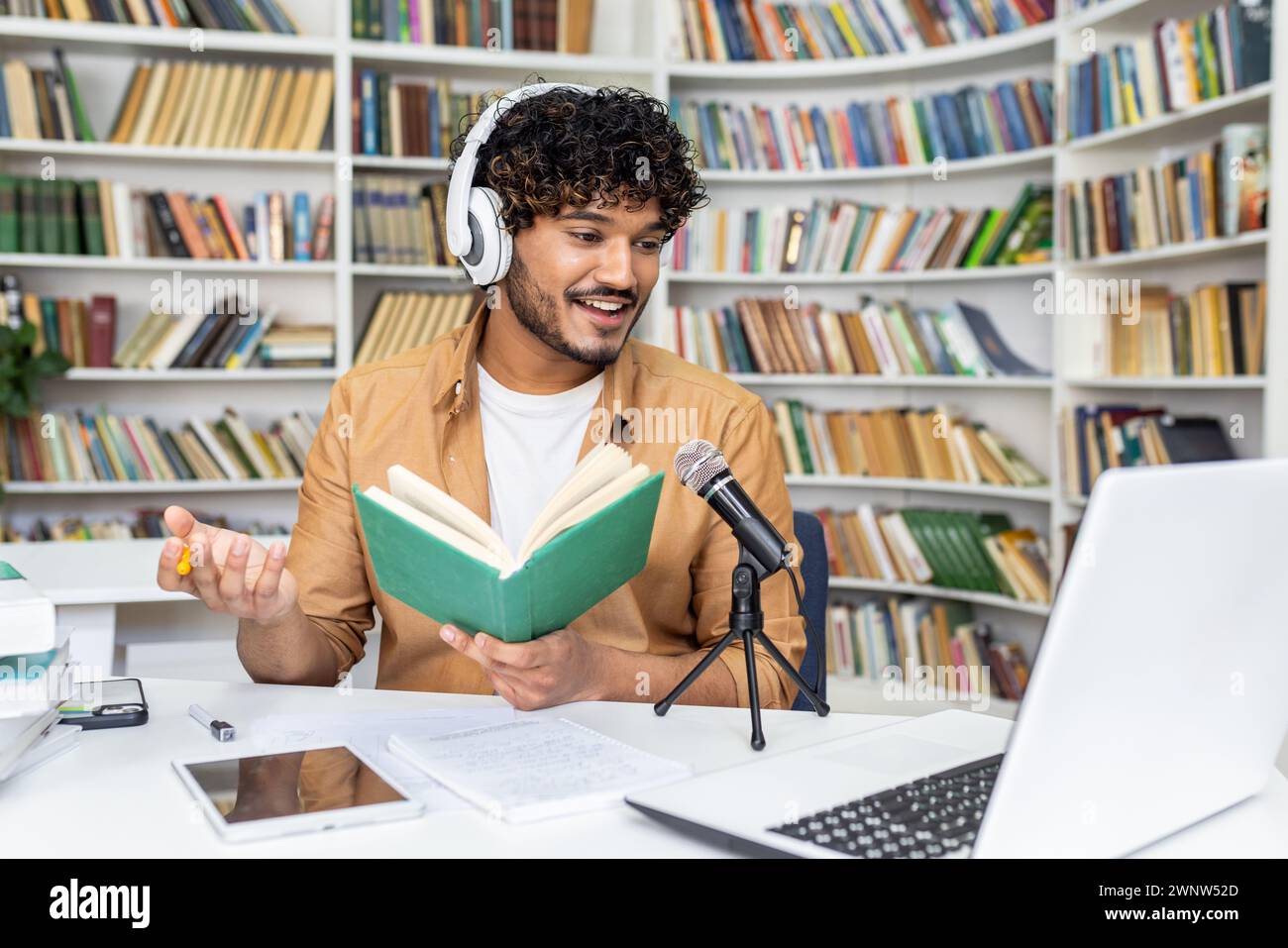 Un giovane podcaster maschile registra in un accogliente studio domestico sullo sfondo di una libreria, evocando un'atmosfera di conoscenza e creatività. Il suo coinvolgimento con il pubblico è palpabile anche attraverso il mezzo visivo. Foto Stock
