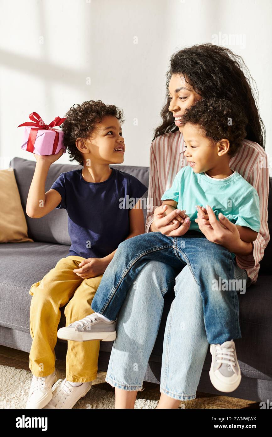 Allegra donna afroamericana che tiene in braccio suo figlio e riceve un regalo da un altro figlio, la festa della mamma Foto Stock