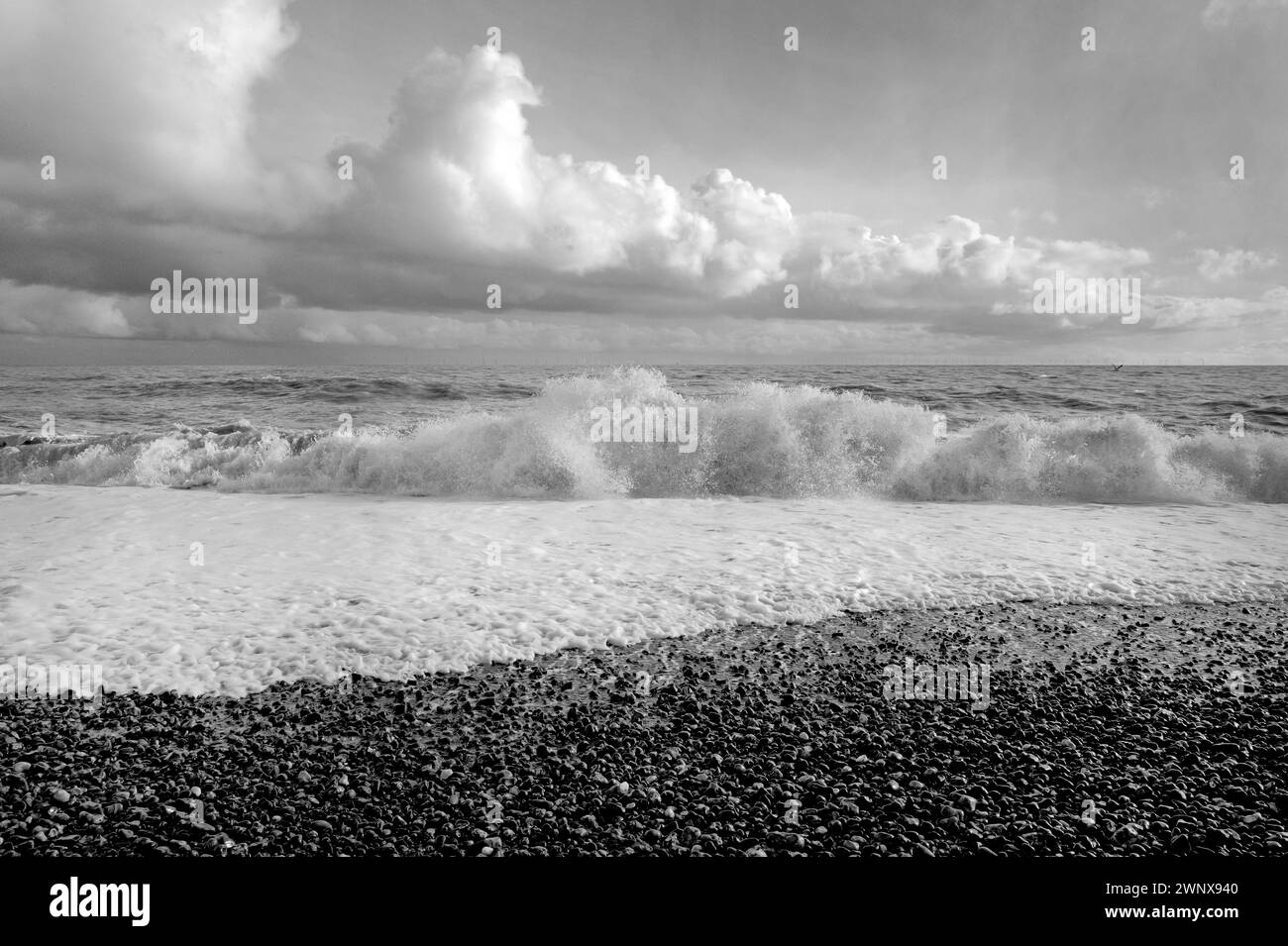 A B&W luce solare forte di onde che si infrangono sulla spiaggia di ciottoli, spruzzi e spruzzi, mare verde, cielo blu con nuvole bianche, costa meridionale, Inghilterra Foto Stock