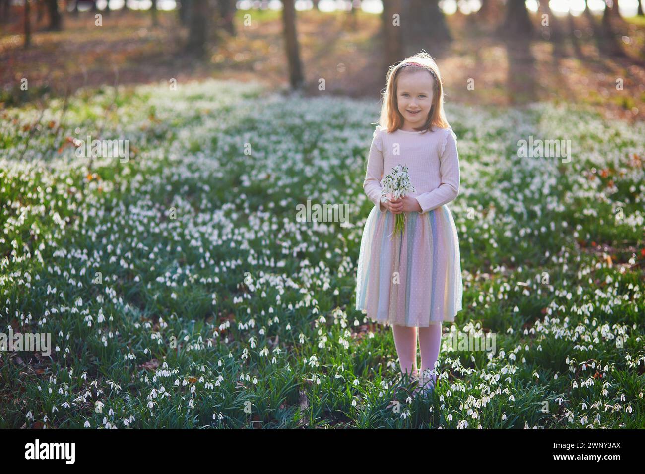 Adorabile ragazza in età prescolare in tutù verde che raccoglie fiori di neve nel parco o nella foresta in un giorno di primavera. Foto Stock