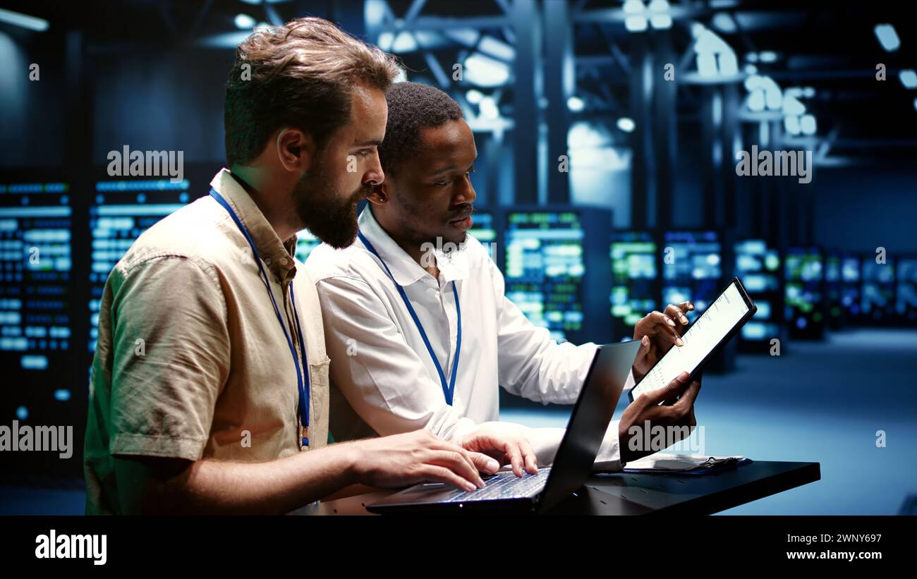 Colleghi che eseguono test diagnostici hardware per data center per determinare e risolvere i problemi software. I tecnici che utilizzano un computer portatile e un tablet per riparare i sistemi critici, controllando la presenza di segnali acustici Foto Stock