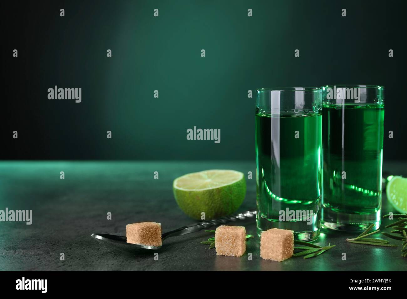 Absinthe in bicchieri da shot, cucchiaio, zucchero di canna, lime e rosmarino su un tavolo testurizzato grigio su sfondo verde, spazio per il testo. Bevanda alcolica Foto Stock