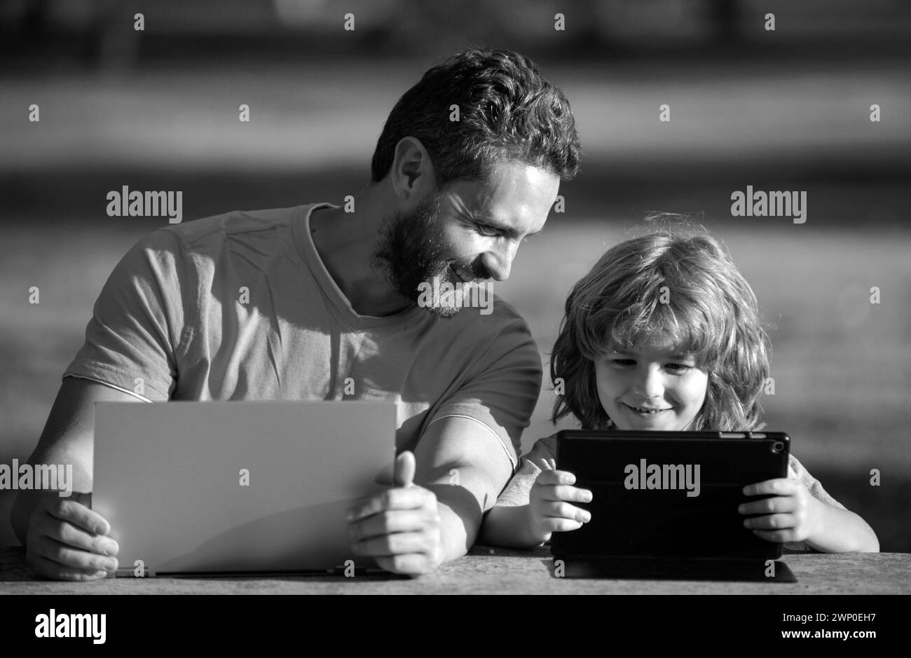 Padre insegna al figlio a usare il laptop, il papà e il bambino della scuola guardando lo schermo del computer e il tablet, giocando, guardando il video, seduto sull'erba Foto Stock