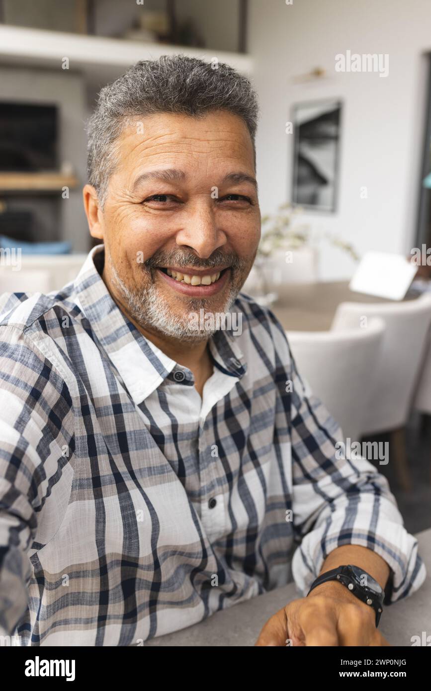 Un anziano birazziale con i capelli grigi sorride calorosamente durante una videochiamata, indossando una camicia a quadri e un orologio Foto Stock