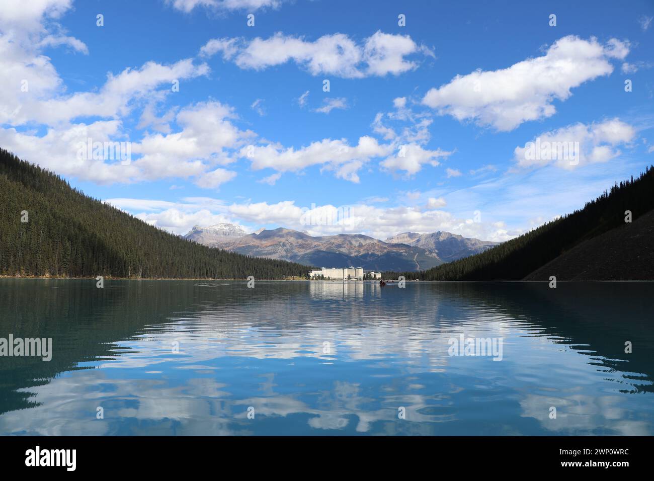 Un'immagine che si affaccia su un hotel annidato alla fine del lago, tra le montagne con acqua calma, foresta verde e montagne in lontananza. Foto Stock